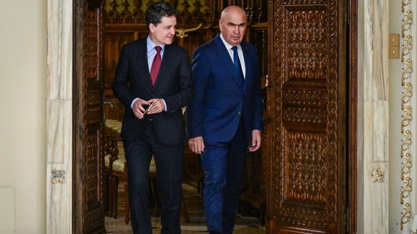 Nicușor Dan și Ilie Bolojan, ambii îmbrăcați în costume elegante, ieșind împreună dintr-o încăpere printr-o ușă masivă din lemn sculptat.