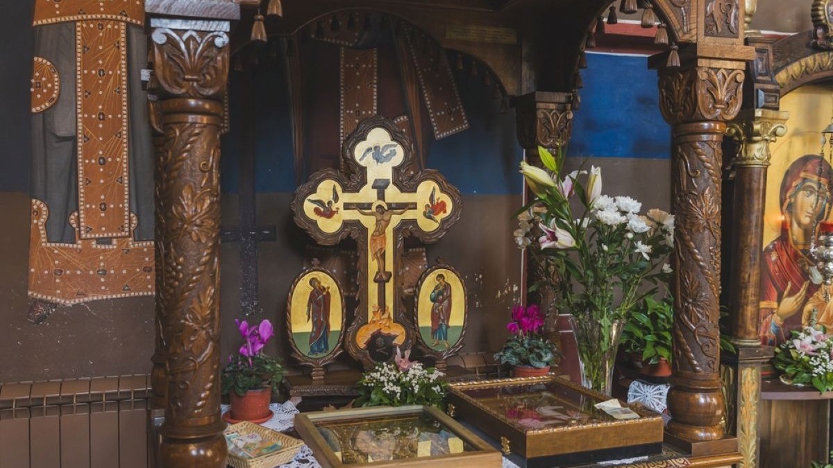 Cruce ortodoxă din lemn sculptat și pictat, așezată pe un altar decorat cu flori naturale într-o biserică.