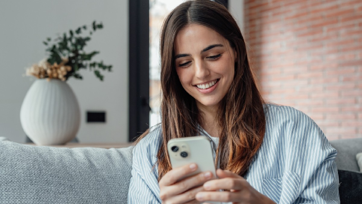 Femeie zâmbitoare uitându-se la ecranul smartphone-ului, așezată pe canapea