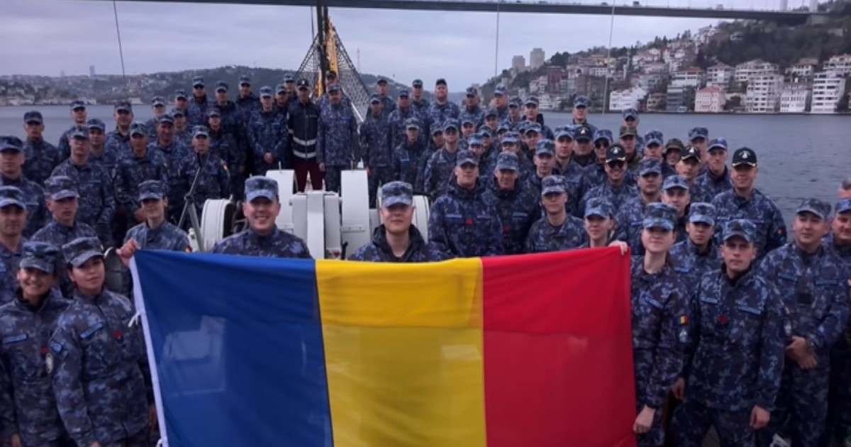 Un grup numeros de militari în uniformă stă aliniat pe puntea unei nave, ținând în față un drapel mare al României. În fundal se vede strâmtoarea Bosfor și orașul, sugerând momentul trecerii navei într-o etapă importantă a călătoriei.