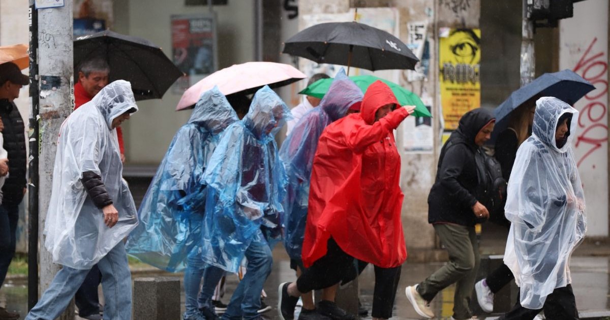 Mai mulți oameni acoperiți cu pelerine de ploaie și umbrele merg pe stradă în zona Piața Romană din București