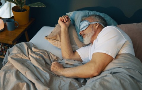 Om senior odihnindu-se în pat cu mască de somn, care apare relaxat și confortabil, înconjurat de perne și pături în dormitor confortabil