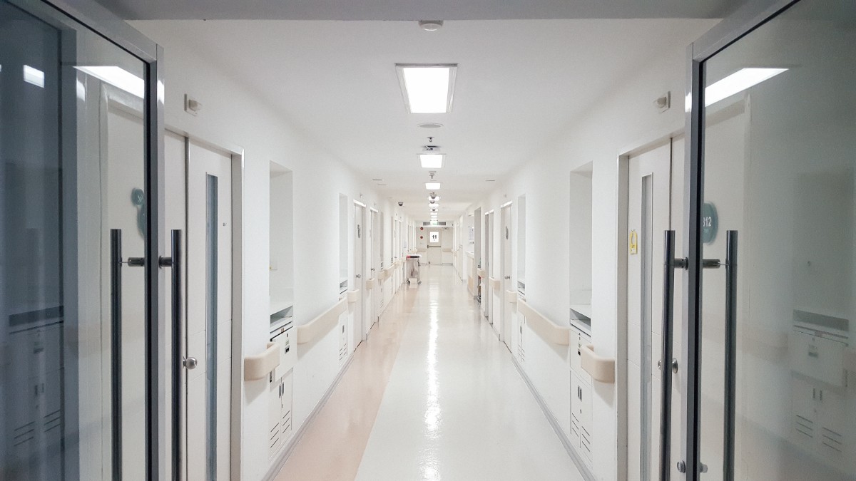 Imaginea surprinde un hol lung și luminos dintr-un spital, cu uși aliniate de-o parte și de alta și iluminare artificială uniformă. Spațiul este curat și ordonat, sugerând un mediu medical modern și lipsit de prezență umană în acel moment.