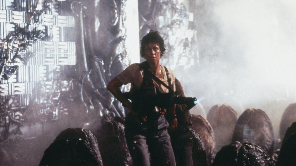Această imagine o prezintă pe Sigourney Weaver în rolul lui Ellen Ripley din filmul Aliens. Ripley apare într-o postură alertă, înarmată cu o pușcă
