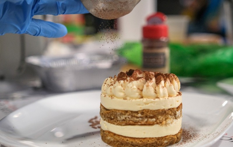 Tiramisu World Cup, un tiramisu construit sub forma de rotund, pe o farfurie. O persoană pune praf de cacao peste el