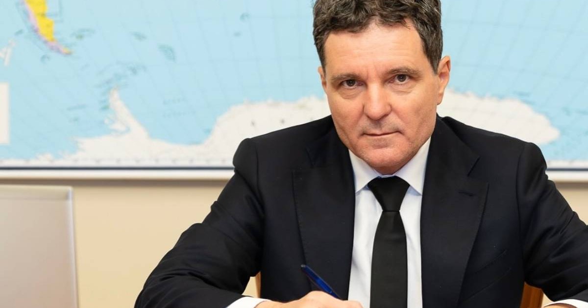 Nicușor Dan, președintele României, în timp ce stă la birou cu pixul în mână, în spatele său are harta lumii și poartă un costum negru