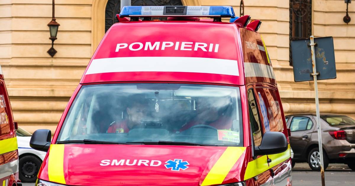 O ambulanță roșie SMURD parcată cu spatele, la volan și pe locul din dreapta al șoferului se află doi asistenți medicali care verifică niște acte