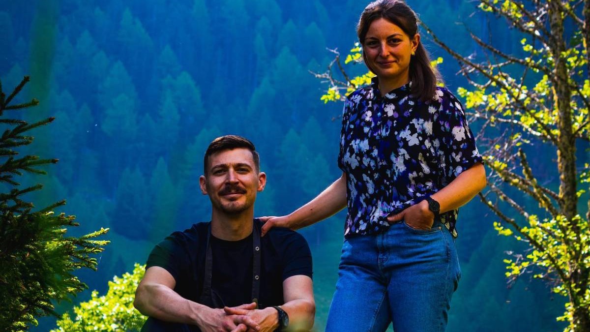 În imagine bucătarul-șef Adrien Lopez și soția lui, Clara, care conduc restaurantul elvetian Au vieux Nendaz , el, in bluza neagra, sta asezat si cu mainile impreunate peste genunchi, ea, cu o bluza albastra cu model floral, sta in picioare, langa el, cu mana stanga bagata in buzunarul din fata de la blugi si mana dreapta pe umarul chefului, in spate un fundal de munte, cu brazi, in umbra