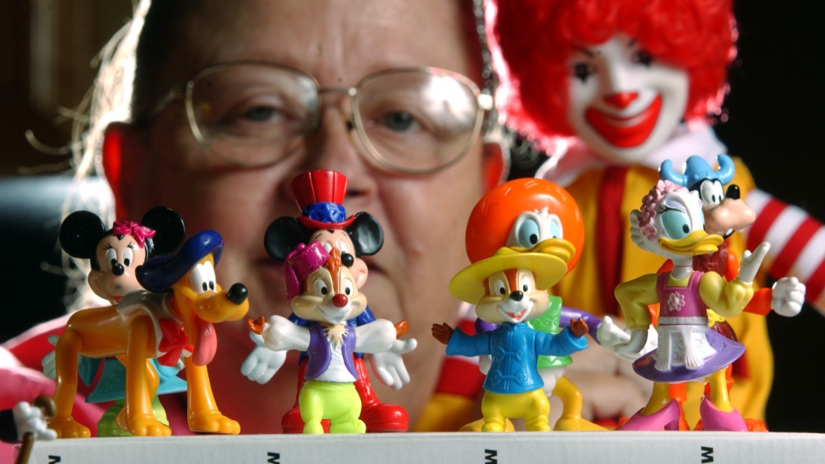 Cinci jucării de la McDonald's, cu figurine Disney, așezate pe o măsuță. În spatele lor se vede o femeie cu ochelari