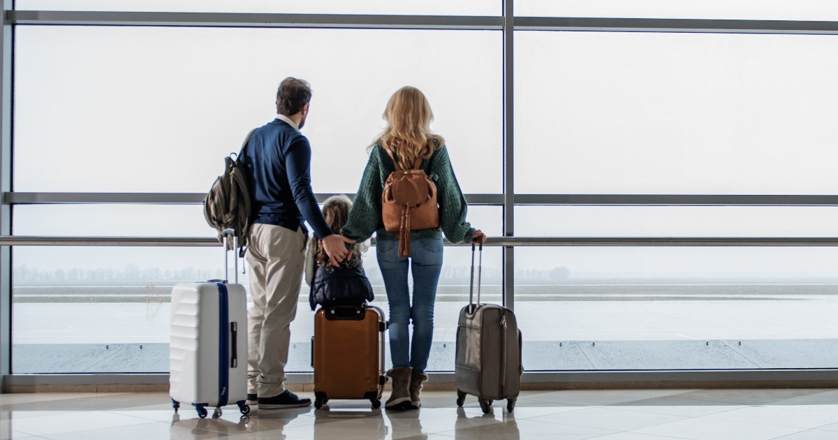 O familie (doi părinți și un copil) stă în fața unui geam mare din aeroport, privind pe geam, pregătită de călătorie. Aceștia sunt înconjurați de bagaje de tip troller.