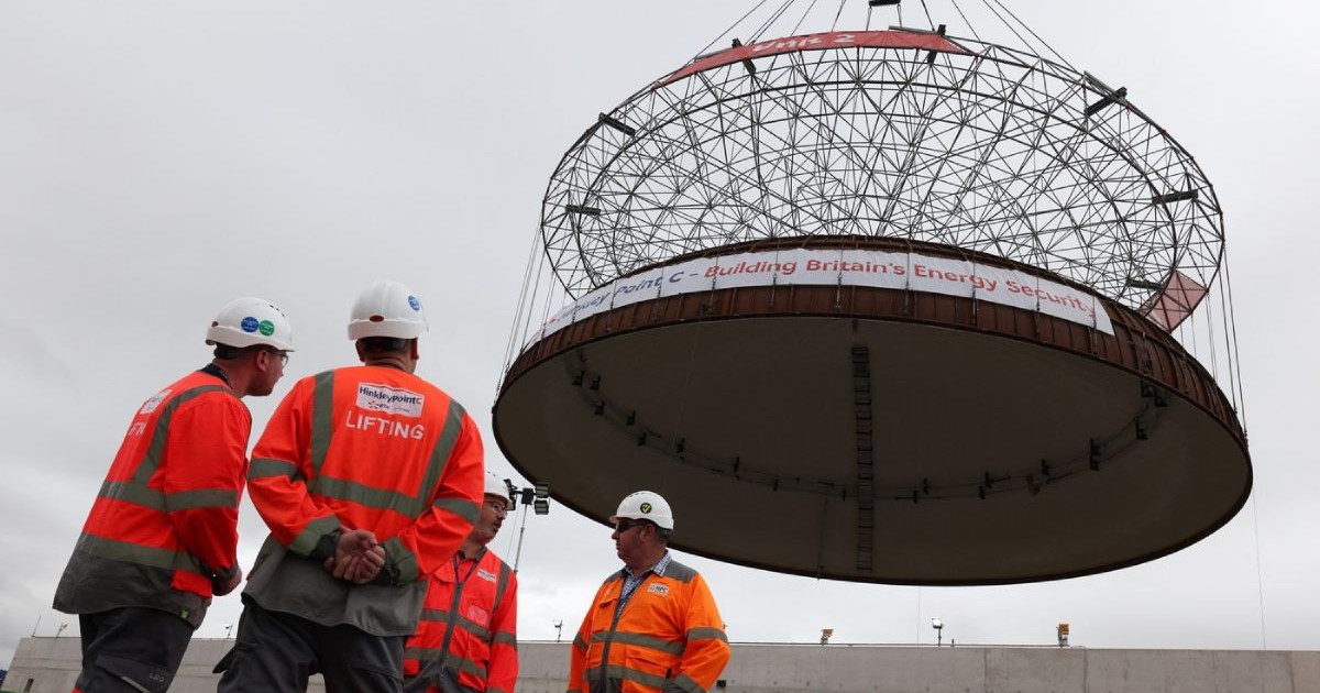 O echipa de muncitori, in costume portocalii, langa mai mare macara din lume - Big Carl - aparent stau sub cupolă de oțel de 245 de tone pe clădirea celui de-al doilea reactor al centralei nucleare Hinkley Point C, pe șantierul de construcție al centralei nucleare din Bridgwater, Somerset. Cupola închide clădirea, permițând instalarea reactorului nuclear și a echipamentului. Data fotografiei: joi, 17 iulie 2025.