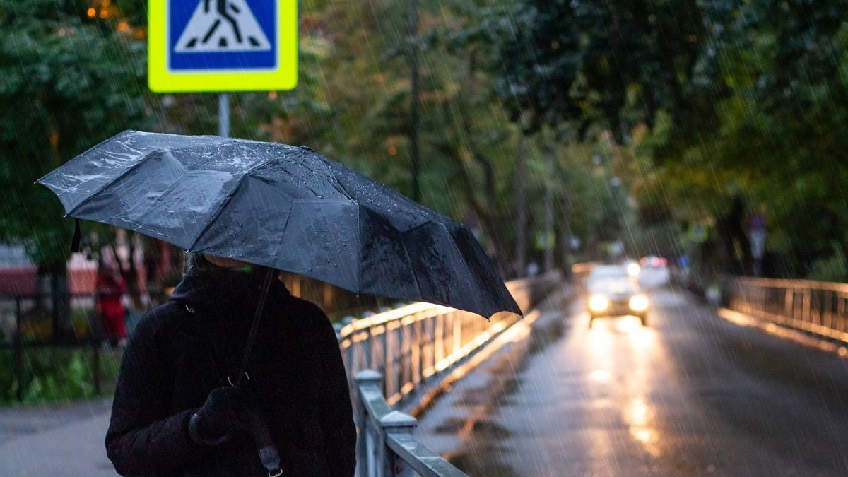 O femeie ține o umbrelă neagră, în timp ce merge pe stradă, iar afară plouă și e frig, iar o mașină cu farurile aprinse vine din față