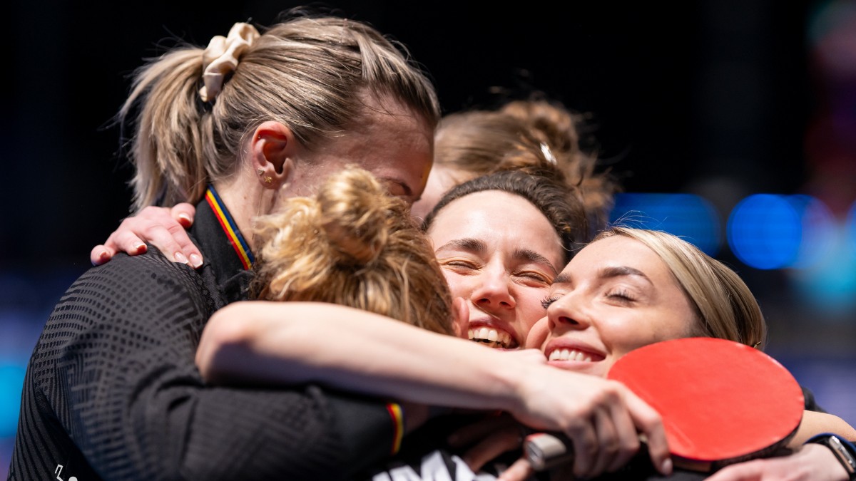 Imaginea surprinde jucătoarele echipei feminine de tenis de masă a României într-un moment de bucurie și emoție după o victorie importantă. Sportivele se îmbrățișează pe teren, celebrând performanța obținută la Campionatul Mondial.
