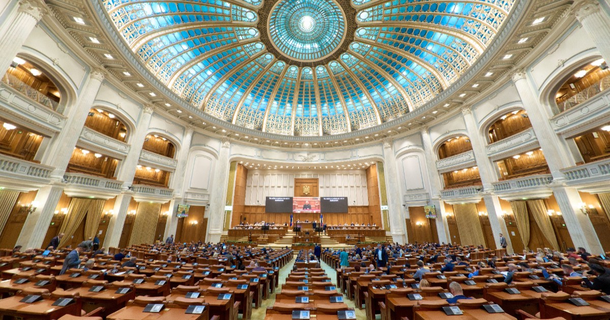 Imaginea prezintă o vedere de ansamblu din interiorul plenului Camerei Deputaților din Palatul Parlamentului, cu un vitraliu albastru pe cupolă și băncile parlamentarilor pregătite pentru ședință.