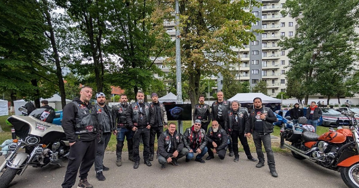 O gască de motocicliști, îmbrăcați în negru, pozează în fața unui parc, cu două motociclete alătuir, una portocalie în dreapta și una argintie în stânga