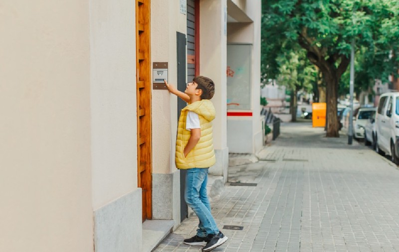 Un băiețel îmbrăcat cu o vestă galbenă și blugi stă pe un trotuar din oraș, întinzându-se pentru a apăsa soneria de la intrarea într-o clădire. În fundal, strada este mărginită de copaci și mașini parcate
