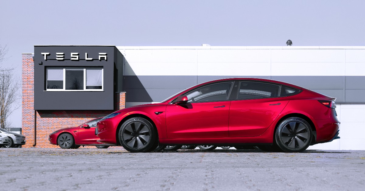 Imaginea prezintă două automobile Tesla, dintre care un model roșu vibrant se află în prim-plan, parcate în fața unei clădiri moderne, cu un semn mare pe care scrie "TESLA". Clădirea are o fațadă simplă, cu secțiuni de cărămidă închisă la culoare și panouri albe, completând estetica modernă a vehiculelor electrice.