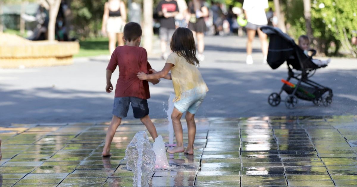 Doi copii, fotografiați cu spatele, se joacă în parc la o fântână de apă la sol, iar în jurul lor se văd oameni și un copil mic în căruț, București, 16 septembrie 2025