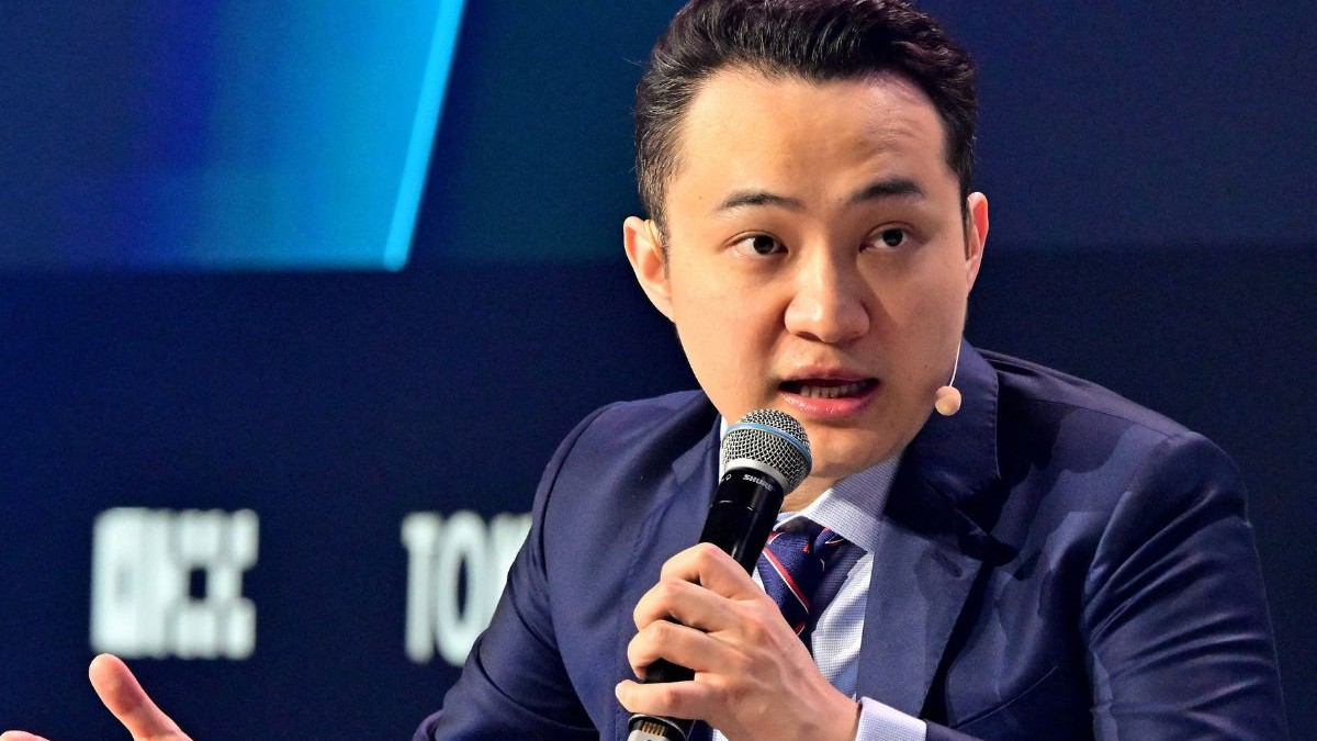 Justin Sun, îmbrăcat cu un costum bleumarin, o cămașă bleu și o cravată bleumarin cu o dungă roșie, ținând un micofon aproape de gură și având sprâncenele ridicate, părând ușor mirat, la Conferința Token 2049 despre criptomonede, la Dubai, pe 1 mai 2025.