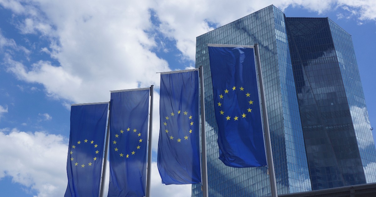 Patru steaguri ale Uniunii Europene flutură pe catarge înalte în fața unei clădiri moderne, compuse din turnuri acoperite cu sticlă de culoare închisă. Steagurile de culoare albastru intens, cu cercul de stele aurii, se profilează pe un fundal de cer albastru cu nori albi pufoși.