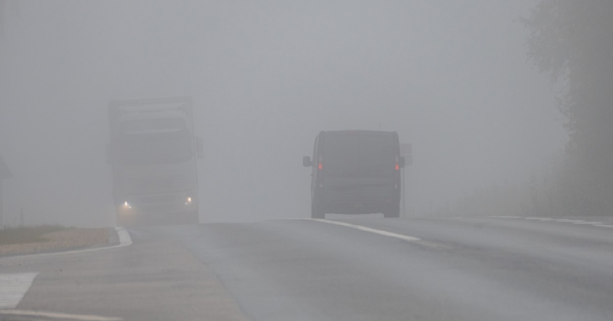 Imaginea surprinde o șosea, cel mai probabil dimineața, acoperită de o ceață densă. Vizibilitatea este redusă, iar drumul este ud. Din cauza condițiilor meteo, contururile obiectelor și ale vehiculelor din fundal sunt neclare. În prim-plan, se văd două vehicule: un autocamion mare, alb, cu farurile aprinse, care se apropie din partea stângă, și o autoutilitară neagră, aflată în fața lui. Ambele par să se deplaseze cu viteză redusă din cauza ceții.