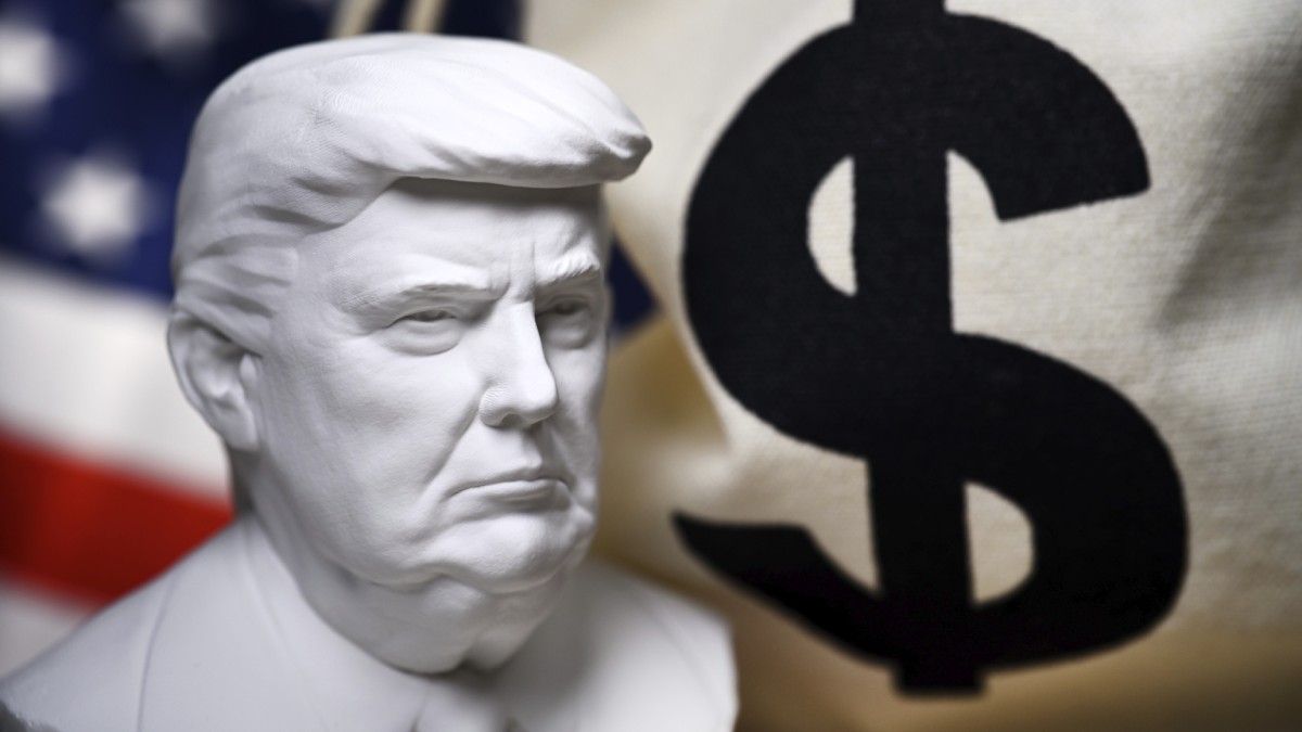În stânga imaginii, o statuetă bust cu Donald Trump. În dreapta, un sac cu semnul dolarului pe el. În spate, stânga, steagul Americii.
