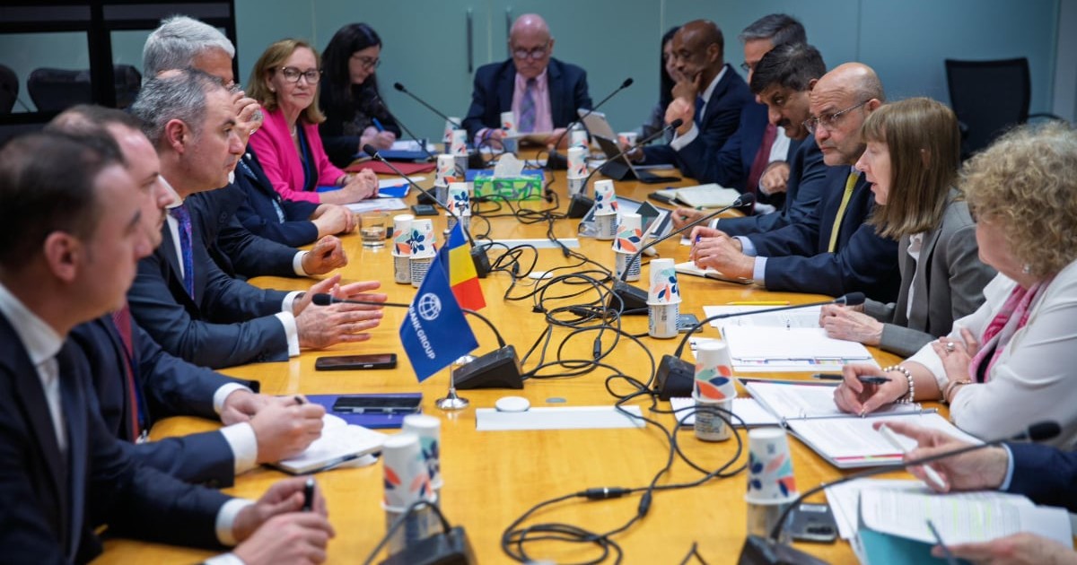 Mai mulți oficiali stau la o masă de negocieri, discutând în cadrul unei întâlniri formale între delegații. Pe masă se află documente, microfoane și steaguri, sugerând o discuție oficială pe teme economice sau politice.