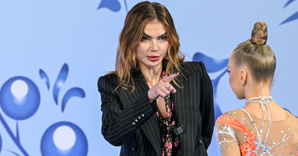Alina Kabaeva îmbrăcată într-un costum negru discută cu o gimnastă care este cu spatele.