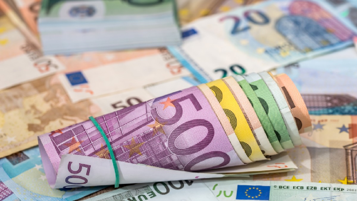 Imaginea prezintă un sul de bancnote euro multicolore prins cu un elastic, așezat peste un fundal format din numeroase alte bancnote de diferite valori, evidențiind în prim-plan cupiura de 500 de euro.