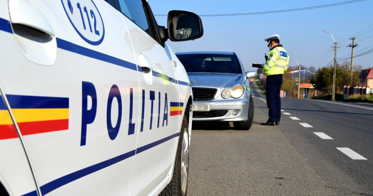 politist aflat langa o masina gri in timp ce scrie, posibil un avertisment sau o amenda. in partea stanga se observa masina politiei