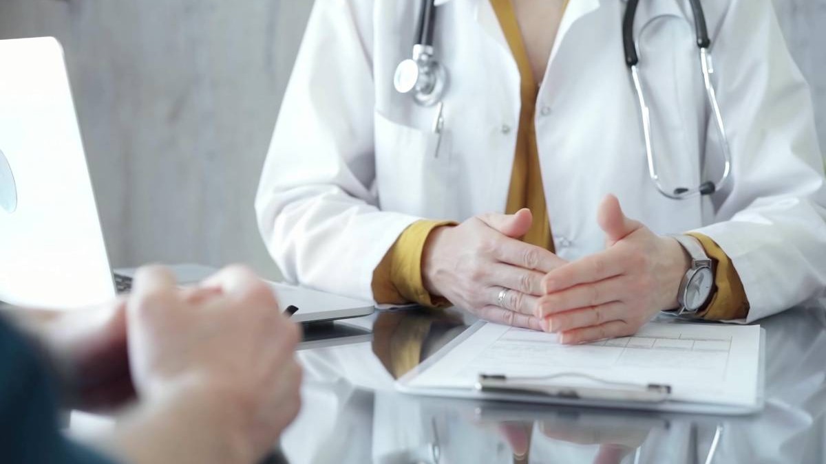 Medic îmbrăcat în halat alb, purtând un stetoscop în jurul gâtului și un ceas de mână, gesticulând ușor deasupra unei mese pe care se află o fișă medicală și un laptop deschis.