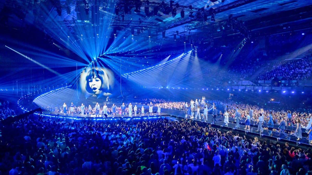 Imaginea surprinde atmosfera spectaculoasă a unui show Eurovision, cu scenă luminată intens și mii de spectatori prezenți în arenă. Efectele vizuale și energia publicului evidențiază amploarea unuia dintre cele mai urmărite concursuri muzicale din lume.