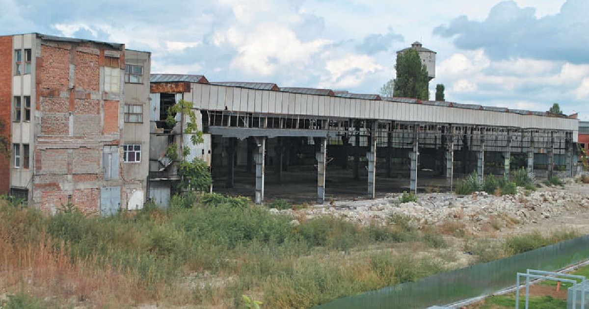 Imaginea prezintă o clădire din cărămidă, în stare de degradare, cu ferestre mari și goale și acoperișul parțial prăbușit, înconjurată de iarbă înaltă și moloz