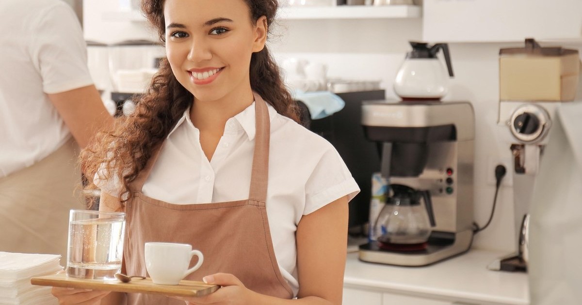 O femeie tânără, cu tricou alb și sorț bej, duce o tava de lemn în mână, pe care are o ceașcă de cafea și un pahar de apa, într-o cafenea