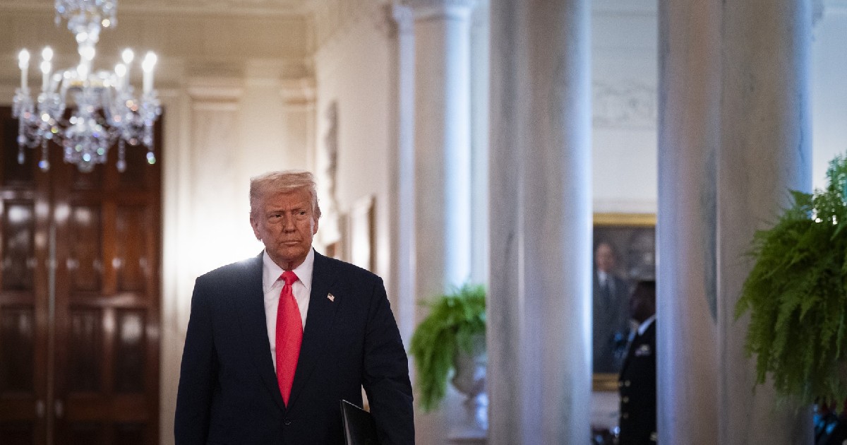 Donald Trump, îmbrăcat într-un costum albastru închis, cămașă albă și cravată roșie, mergând pe un hol. În dreapta lui sunt niste ferestre cu perdele albe, în stânga sus este un candelabru.