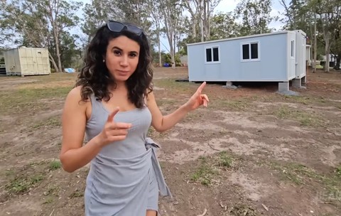 O femeie îmbrăcată într-o rochie gri care arată spre o casă modulară prefabricată, de culoare deschisă, amplasată într-un spațiu exterior deschis.