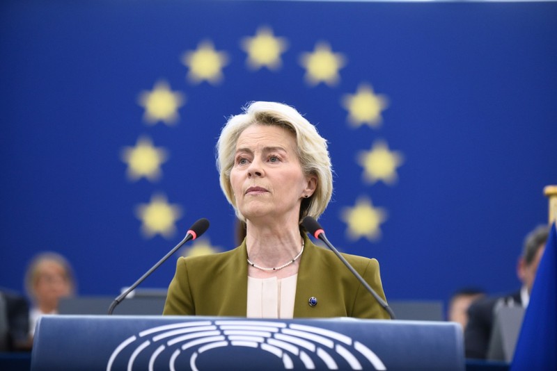 Imaginea o arată pe Ursula von der Leyen vorbind la un pupitru. Are părul scurt, blond și poartă un sacou verde-măsliniu peste un top de culoare crem. Un colier de perle este vizibil la gât. Fundalul este un steag albastru cu un cerc de stele galbene. Două microfoane se află în fața ei Foto: Profimedia