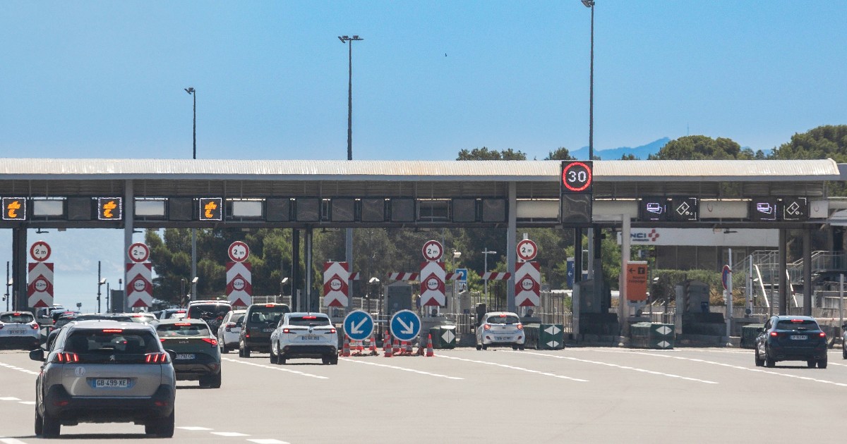 Imaginea prezintă mai m ulte mașini care se apropie sau staționează la barierele de taxare pe o autostradă, cu mai multe benzi ide circulație