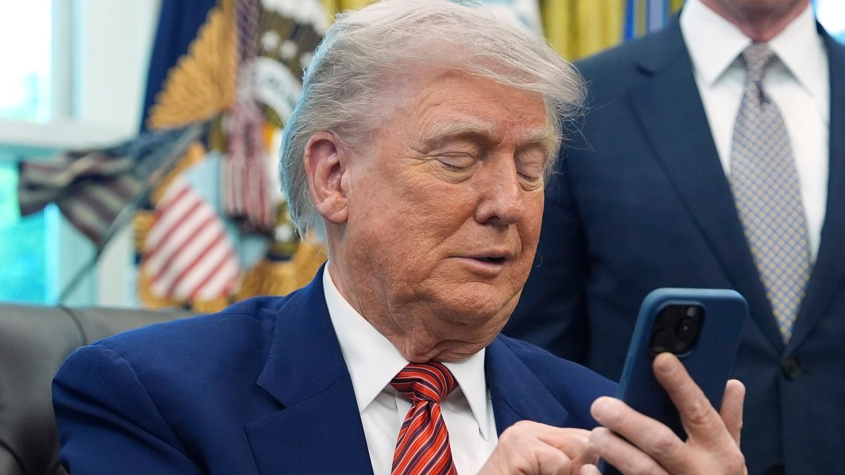 Donald Trump surprins în timp ce ține în mâini un telefon. Președintele american poartă un sacou albastru, o cămașă albă și o cravată roșie cu dungi negre