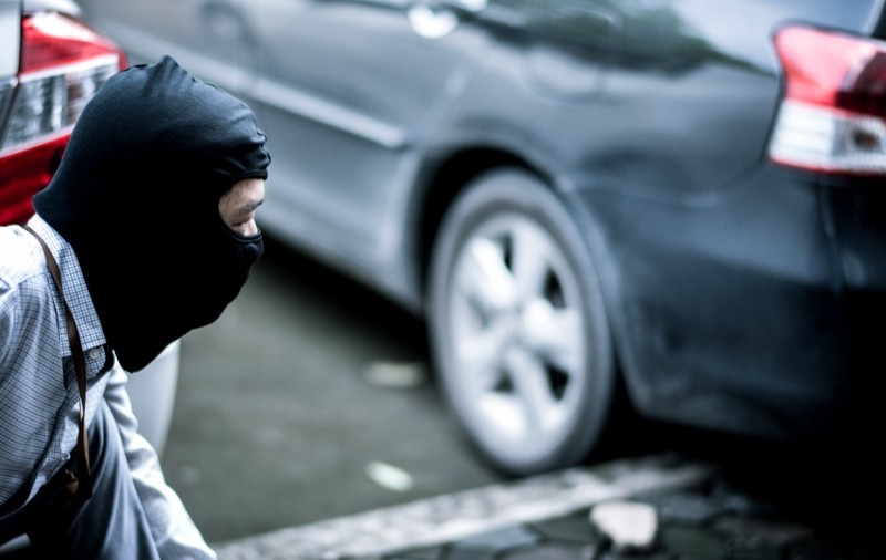 Un bărbat purtând o cagulă neagră, ghemuit discret lângă un autoturism parcat.   