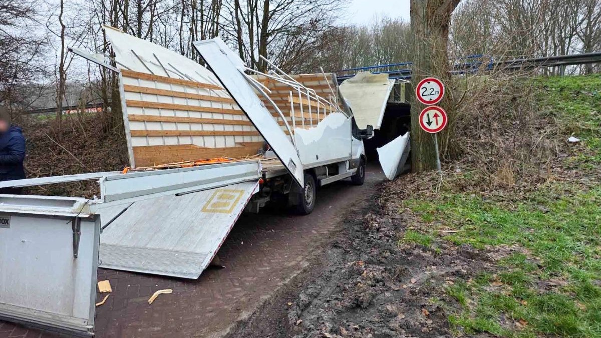 Un camion alb cu remorcă este blocat într-un viaduct, pe un drum îngust și noroios, cu iarbă verde, copaci și un stâlp pe care se află două indicatoare.