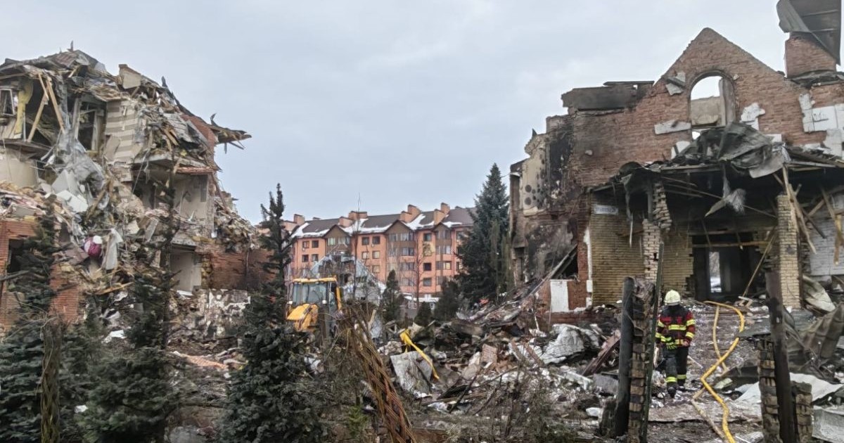 o scenă de distrugeri masive în exterior, unde două clădiri din cărămidă, situate în stânga și în dreapta cadrului, sunt parțial prăbușite, lăsând la vedere structurile interioare de lemn și beton. Spațiul dintre ele este acoperit de un morman mare de moloz compus din resturi de zidărie, grinzi rupte și bucăți de izolație, iar în centrul acestui perimetru se află un buldoexcavator galben parțial vizibil, Borșceahivka, 23 februarie 2026