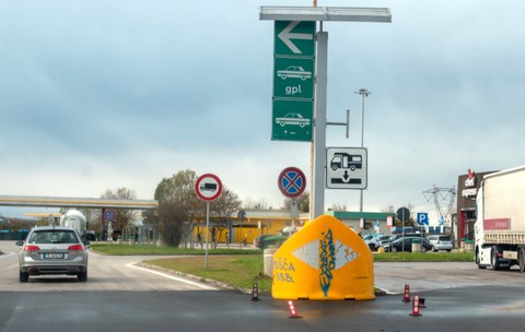 O zonă de servicii și stație de alimentare de pe o autostradă din Italia, cu panouri verzi de direcționare, indicatoare rutiere, un autoturism Volkswagen argintiu în mers și un camion staționat în dreapta.