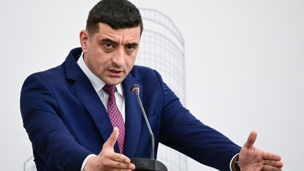În imagine este ilustrat George Simion, îmbrăcat în costum albastru închis și cămașă albă. Acesta vorbește la microfon
