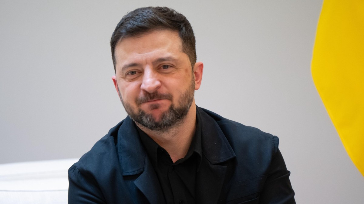 În imagine Volodimir Zelenski, presedintele Ucrainei, îmbrăcat într-un costum închis la culoare, cu barbă scurtă și păr tuns scurt, așezat într-un decor sobru. În fundal se distinge parțial un steag galben, iar expresia sa este calmă și concentrată, sugerând un cadru oficial.