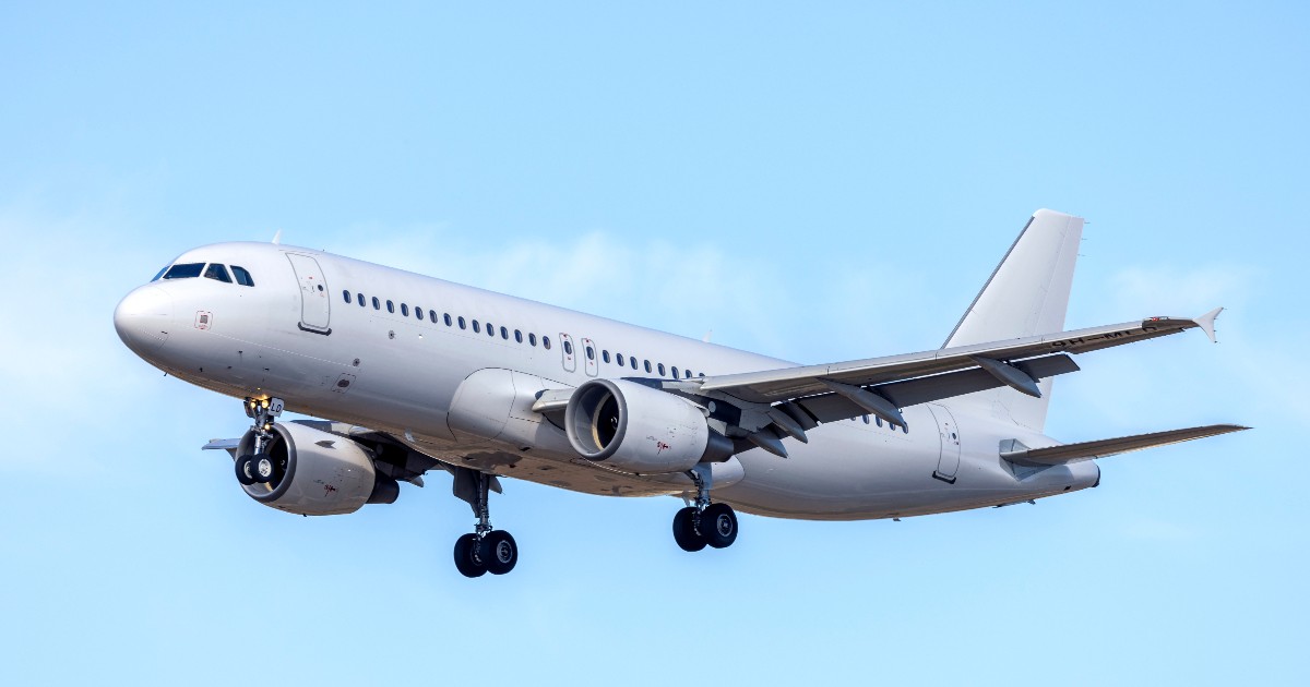 Un avion de pasageri cu fuzelaj de culoare gri-pal, având trenul de aterizare coborât, este surprins în zbor pe un fundal de cer albastru deschis, probabil în timpul manevrei de aterizare sau decolare. Aeronava, care pare a fi un model din familia Airbus A320 (sau similar), este echipată cu două motoare cu reacție montate sub aripi și prezintă un șir de hublouri mici de-a lungul cabinei.