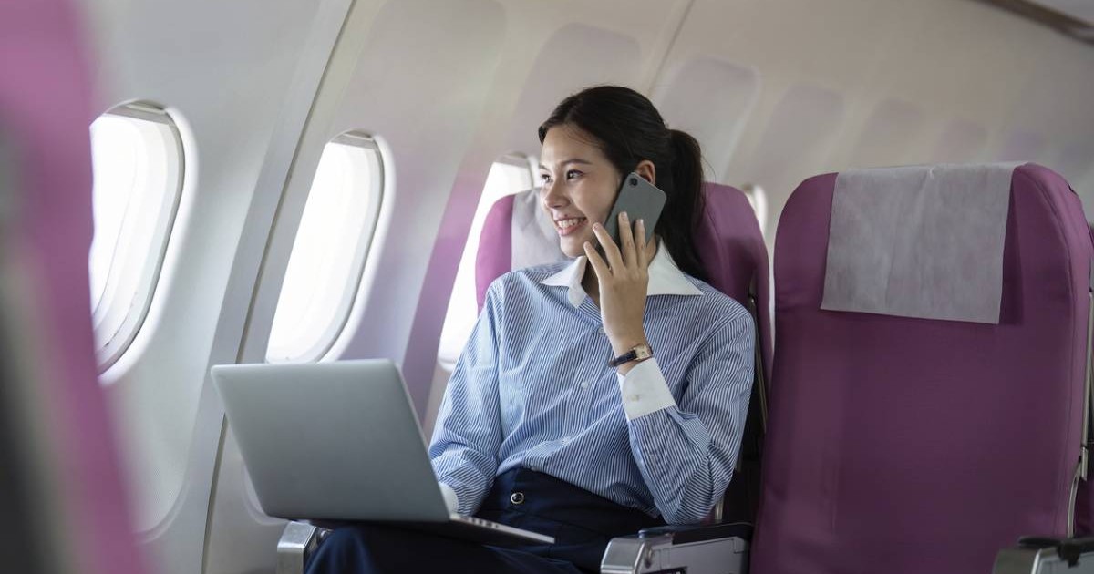 O femeie zambeste si se uita pe fereasta in timp ce folosește un laptop și vorbește la telefon , așezată într-un avion, scaunele sunt mov