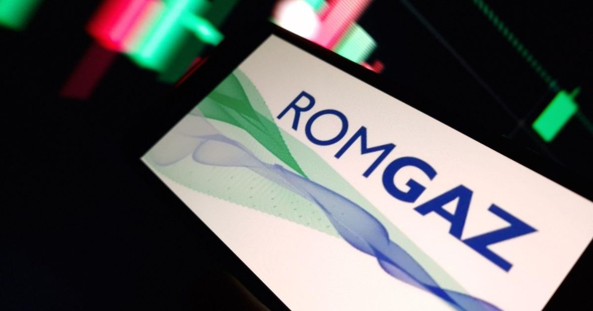 Sigla companiei Romgaz afișată pe ecranul unui telefon mobil, într-o camera întunecată cu alte ecrane pe care sunt afișate cotații bursiere colorate în roșu și verde