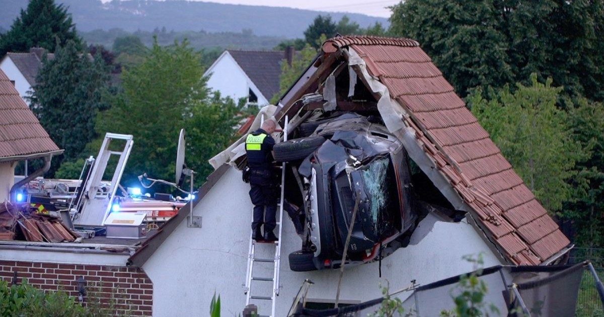 In imagine se vede o masina blocata intr-un hambar al unei case, langa ea un pompier urcat pe o scara, in spate se vad masini de interventie cu semnale luminoase pornite albastre-rosii. 19 iulie 2025, Germani