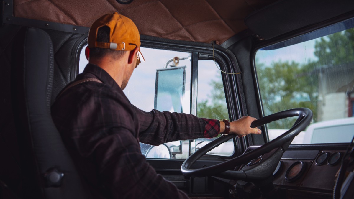 Imaginea prezintă un șofer purtând șapcă aflat la volanul unui camion, privind spre oglinda laterală în timp ce conduce.